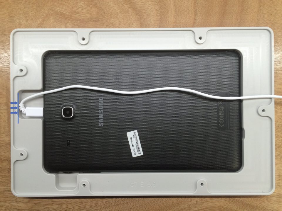 Samsung tablet with black back panel connected to a white charging cable in a plastic frame on a wooden surface